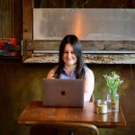 Copywriter Alex Haynes sitting at a cafe table, working on a laptop in front of her.